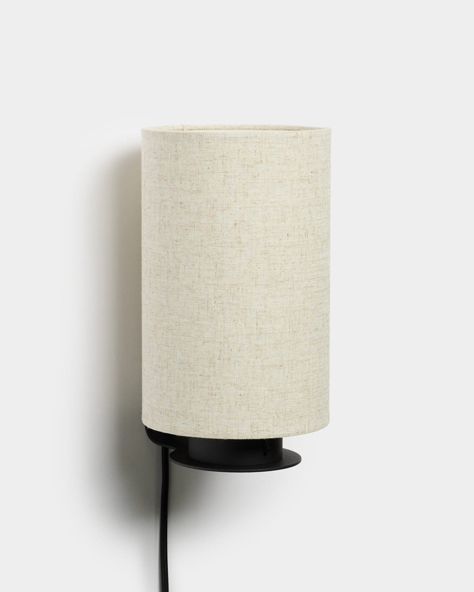 Feya wall lamp by WoodUpp with a black metal base, textured linen-cotton shade in beige, and an exposed black power cord. Nordic minimalist design.